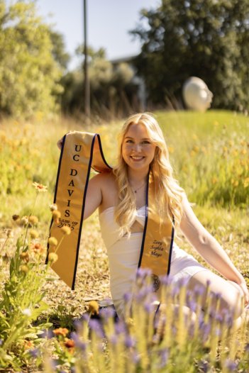 A young white blonde woman sits in a small meadow of flowers wearing a white dress and UC Davis graduation stole smiles and holds up "UC Davis" part of stole to the camera.